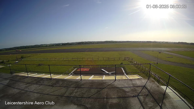 A clear view of an airfield on a sunny day, with a grassy area, runways, and a small helipad with a white helicopter silhouette. There is a wooden fence and a flagpole with a flag on the left side. The sky is mostly clear with the sun shining brightly, and trees are visible in the distance. The timestamp shows "01-06-2025 08:00:23," and the location is indicated as "Leicestershire Aero Club."