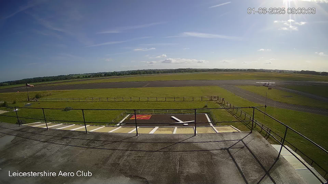 A wide view of an airfield showing a small airplane on the runway, green fields stretching into the distance, a windsock on the left, and a bright blue sky with some scattered clouds. The timestamp in the upper right corner reads "01-06-2025 09:00:03," and at the bottom left, text indicates the location as "Leicestershire Aero Club."