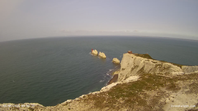 An aerial view of white chalk cliffs extending into the sea, with a small structure on top of the cliff. In the distance, a lighthouse stands on rocks in the water next to the cliff. The sky is clear with a slight haze near the horizon. The timestamp in the bottom left corner reads "01/06/2025 09:00:31," and the website "isleofwight.com" is visible in the bottom right corner.