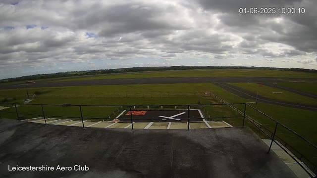 An empty aircraft hangar with a helipad on the ground. Outside the hangar, there is a grassy area with a windsock and a flagpole. The scene includes a runway stretching into the distance under a cloudy sky. The timestamp shows "01-06-2025 10:00:10," and the bottom left corner reads "Leicestershire Aero Club."