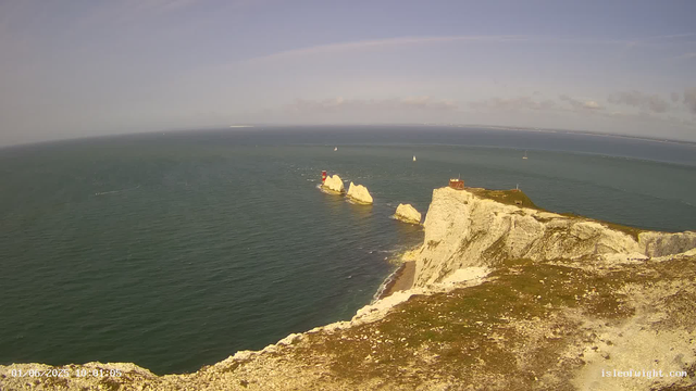 A panoramic view of white chalk cliffs extending into the sea on a clear day, with a small lighthouse and a building on top of the cliffs. The ocean has several sailboats, and the sky is mostly clear with a few wispy clouds. The image appears to be taken from a webcam.