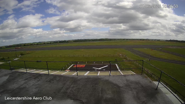 An outdoor view from a webcam at Leicester Aero Club shows a runway stretching into the distance under partly cloudy skies. In the foreground, there is a helipad with a large white "X" marking and a red square with some text, surrounded by a safety railing. The scene is calm and open, with green grassy fields and a line of trees far in the background. The timestamp in the upper right corner indicates the date and time as June 1, 2025, at 10:59:50.