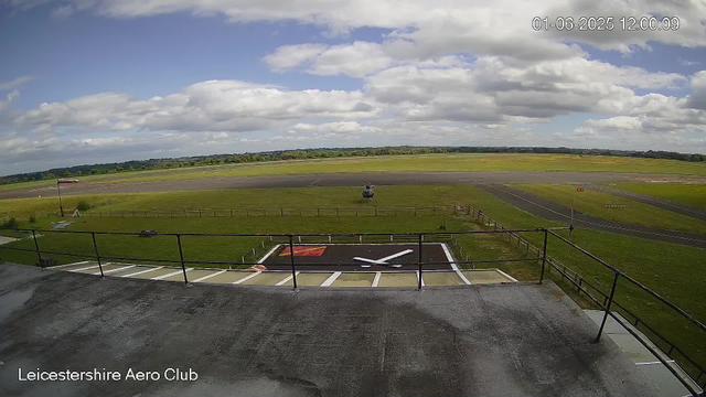 A view from a webcam at Leicesterhire Aero Club showing a grassy airfield with a small helicopter parked on the grass. The sky is partly cloudy with patches of blue, and the horizon stretches far into the distance with many trees visible. The camera is positioned above a parking area with painted markings, and a sign reading "Leicestershire Aero Club" is at the bottom of the image. The timestamp indicates the date as June 1, 2025, and the time as 12:00:09.