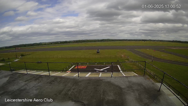 A view from a webcam at Leicestershire Aero Club showing an outdoor helipad with a helicopter parked on a grassy area. In the background, there is a runway stretching across a flat landscape with a partly cloudy sky overhead. The foreground includes a wet concrete platform with a railing, and an area marked with a clock-like design on the ground. The scene appears to be on a slightly overcast day.