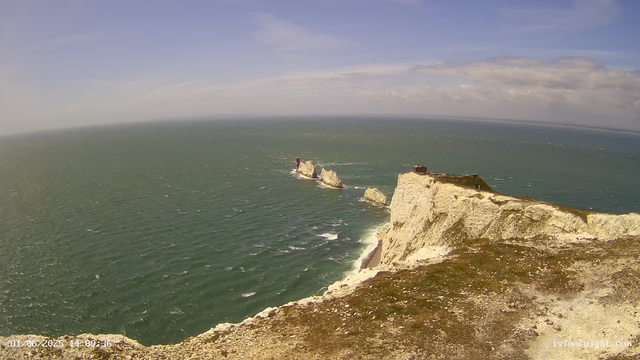 An aerial view of white chalk cliffs overlooking the ocean, with a small structure on top of the cliff. In the distance, several rock formations and arches are visible in the water, with a lighthouse on one of the rocks. The sky is partly cloudy with patches of blue.