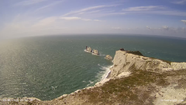 An aerial view of white chalk cliffs overlooking the ocean. The cliffs are topped with grassy terrain, and there are several rock formations and small structures visible near the edge. The sea below has gentle waves, and the sky above is partly cloudy with some wispy clouds. The timestamp on the bottom left reads "01/06/2025 15:00:30".