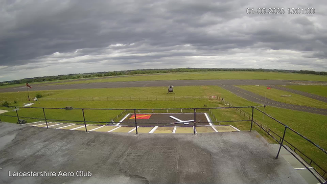 A view from a webcam at Leicestershire Aero Club shows a small helicopter parked on the grass near a helipad with a red outline and white directional arrow. In the background, there is a runway, some wind socks, and safety barriers. The sky is overcast with dark clouds. The image includes a timestamp in the top right corner and the location label "Leicestershire Aero Club" at the bottom left. A helicopter sits on the grass near the runway, and the area appears to be an airfield with open green spaces and paved runway tracks.