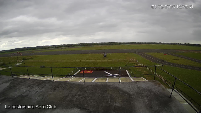 A helicopter is hovering just above the ground on a grassy airfield with a paved runway in the background. The sky is overcast with thick, gray clouds. There is a windsock on the left side of the image, indicating the wind direction. The foreground shows a wet rooftop or balcony with railing, and the bottom left corner displays the text "Leicestershire Aero Club." The timestamp in the top right corner reads "01-06-2025 15:59:51".