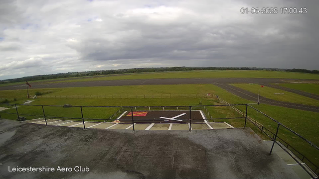 A view from a webcam at Leicestershire Aero Club shows a cloudy sky over an airfield with a runway stretching into the distance. In the foreground, there is a helipad with a large white "H" marking, fenced off with a small staircase leading down to the grass. An anemometer with a wind sock is situated on the left side of the image. The date and time displayed in the upper right corner is 01-06-2025 at 17:00:43.