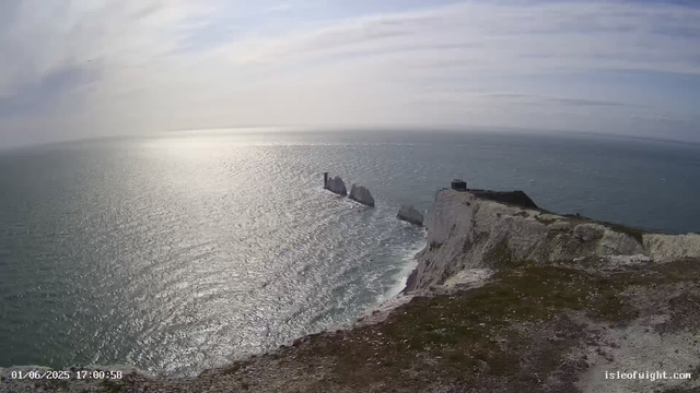 A view from a webcam shows a coastal landscape with white chalk cliffs meeting the ocean. The sea is calm and glistening under the partly cloudy sky, with some large rock formations visible rising from the water near the cliffs. There is a small dark building or structure on top of the cliff in the right part of the image. The scenery appears to be during late afternoon, as indicated by the timestamp.