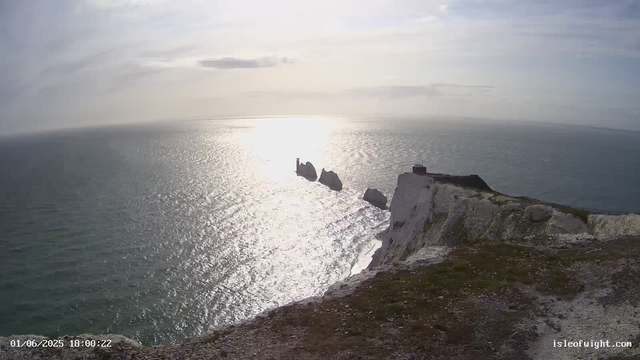 A view of a coastal landscape with white chalk cliffs extending into the ocean. In the distance, there are large sea stacks rising from the water, one of which has a lighthouse on top. The sunlight reflects off the water, creating a bright, shimmering effect. The sky is partly cloudy, and the overall scene appears calm and serene.