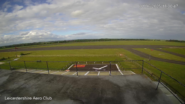An outdoor scene at Leicestershire Aero Club shows a paved balcony or rooftop with a black railing in the foreground. Beyond the railing, there is a grassy area with a marked helicopter landing pad featuring a large white "H" and a red rectangle. The background includes a wide open airfield with several runways, grassy areas, and distant trees under a partly cloudy sky with patches of blue. The timestamp shows the date as June 1, 2025, and the time as 18:00:47.