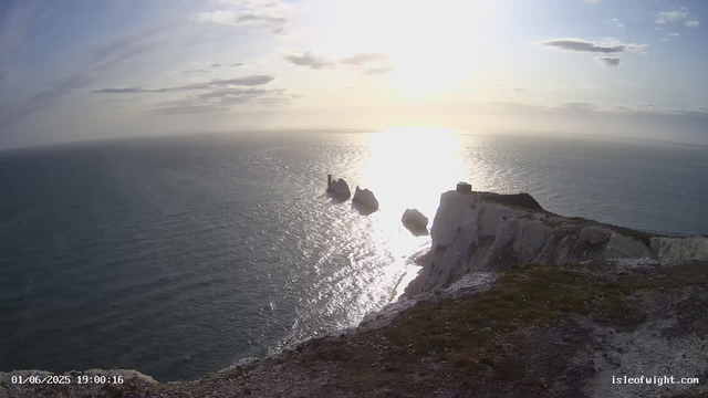 A view of a coastal landscape during sunset, showing a white chalk cliff in the foreground with a grassy top. Below the cliff, the sea extends to the horizon, with the sun reflecting off the water. In the distance, there are rock formations emerging from the sea, including a small lighthouse on one of the rocks. The sky has scattered clouds, and the overall scene is illuminated by the setting sun, creating a bright, serene atmosphere.