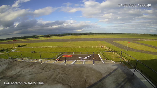 The image shows an outdoor view from a webcam at Leicestershire Aero Club. In the foreground, there is a rooftop with a large clock painted on the surface, showing the time as around 9:40. The middle ground features a grassy area enclosed with a white fence, with a small flagpole flying a wind sock. The background includes a runway extending into the distance, surrounded by open fields and a partly cloudy sky with patches of blue. The time and date displayed on the top right corner are June 1, 2025, at 19:00:48.