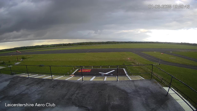 Image of an empty airplane parking area at Leicestershire Aero Club during cloudy weather, with dark storm clouds overhead and green fields in the distance. The timestamp shows the date and time as June 1, 2025, 19:59:46. No aircraft or people are visible.