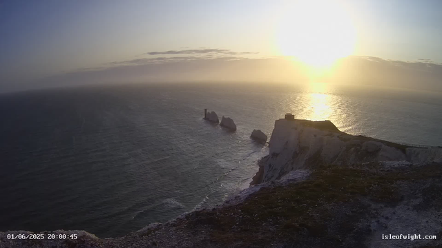A view of the ocean at sunset, with white chalk cliffs in the foreground. The cliffs have some patches of grass, and a small building or structure is visible on top of one of the cliffs. In the distance, there are several large rock formations jutting out of the water, and the sun is low on the horizon, casting a golden glow over the scene. The sky has some scattered clouds. The timestamp shows "01/06/2025 20:00:45" and the website "isleofwight.com."