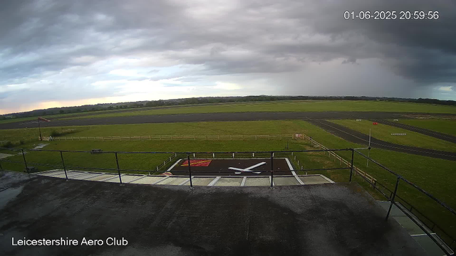 A wide-angle view from a CCTV camera at Leicestershire Aero Club shows an empty airfield under a cloudy sky. The foreground features a helipad with a large white "H" and a red nearby marking, surrounded by a safety railing. Beyond, there is a grassy area with a small windsock on a pole and a fence, small runway strips, and open fields extending to a distant horizon. The sky is partly cloudy with dark storm clouds on the right, and lighter clouds on the left. The date and time display in the upper right corner reads "01-06-2025 20:59:56."