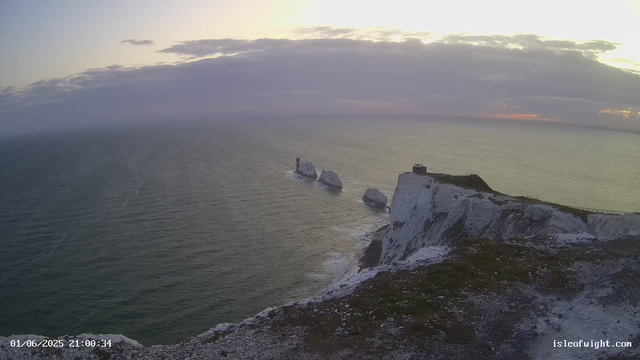 A view of white chalk cliffs along the coastline overlooking the ocean during sunset. The sky is partly cloudy with the sun casting a warm glow, and the sea appears calm with gentle waves. The cliffs extend into the water, with some structures visible on top of the cliffs. There are also notable rock formations rising from the sea near the cliffs.