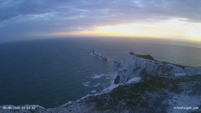 A panoramic view of white chalk cliffs rising from the ocean at sunset. The sky is partly cloudy with hues of purple, pink, and orange near the horizon, and the sea below is calm with gentle waves. A lighthouse stands on one of the cliffs, and a small building is visible on a distant hill. The timestamp shows the date and time as June 1, 2025, at 21:59:42.