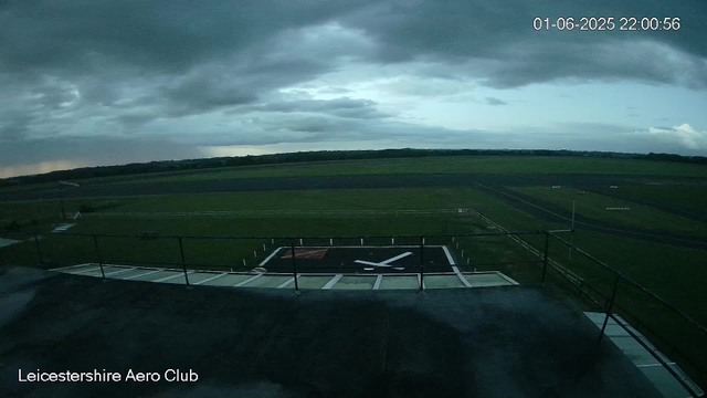 A view from a webcam showing an overcast sky with dark clouds over a grass-covered airfield. In the foreground, there is a helicopter landing pad marked with a large white "H" inside a black square, surrounded by railings. The landscape is flat, with green fields extending into the distance, and a distant tree line on the horizon. The image is labeled "Leicestershire Aero Club" at the bottom left corner, and the date and time displayed in the top right corner is "01-06-2025 22:00:56."