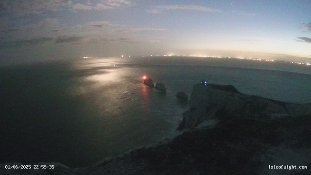 A view of a rocky coastline at dusk or dawn with a calm body of water. There are rocks jutting out into the water, some illuminated by small lights, with the horizon showing a distant shoreline illuminated by many lights. The sky is partly cloudy with a faint glow from the setting or rising sun. The timestamp in the bottom left indicates the date and time as January 6, 2025, at 22:59:35. The website "isleofwight.com" is visible in the bottom right corner.