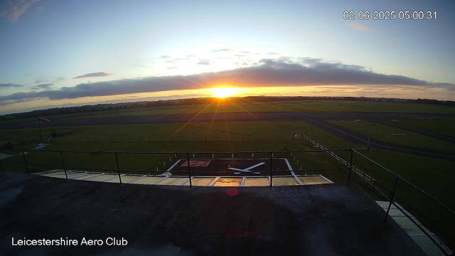 A sunrise over an airfield with a clear view of runways and a helipad at Leicester Aero Club. The sky has some clouds, and the sun is just beginning to rise on the horizon. The image is timestamped February 6, 2025, at 5:00 AM.