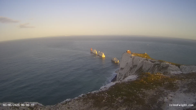 A panoramic view of white chalk cliffs rising sharply from the ocean, with a small building and lighthouse on the top of the closest cliff. Several large chalk stacks are visible offshore, with a lighthouse on the tallest stack. The sky is clear with a few scattered clouds, and the ocean appears calm with gentle waves. Sailboats are visible in the distance. The timestamp in the bottom left corner shows "02/06/2025 06:00:49" and the website "isleofwight.com" is in the bottom right corner.