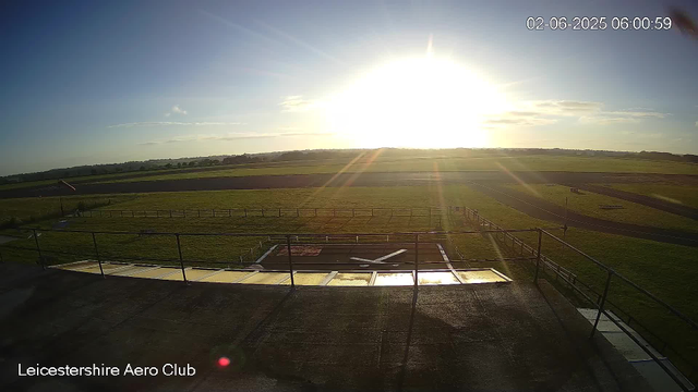 A wide view of an airfield at sunrise or sunset, with the sun low on the horizon casting a bright glare and lens flare. The foreground features a rooftop with a railing and various equipment, while the background shows open grass fields, runways, and a distant horizon with a few scattered clouds. The date and time are displayed in the upper right corner.