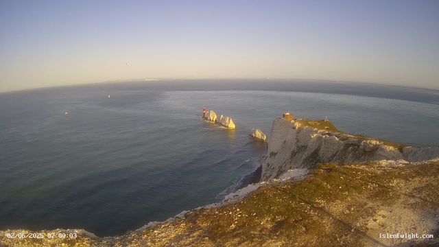 A view from a webcam showing white chalk cliffs with a grassy top overlooking the sea during sunrise or sunset. There is a small building and a lighthouse on the edge of the cliffs, with a few scattered rocks in the water below. The sky is clear with a soft gradient of light. The image includes a timestamp at the bottom left corner reading "02/06/2025 07:00:03" and the website "isleofwight.com" at the bottom right corner.