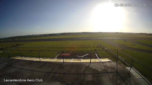 An outdoor view of an airfield at sunrise or sunset, with a clear sky and the sun near the horizon. The foreground shows a concrete balcony with a railing, and a small weather vane or wind direction indicator on the ground. The airfield has grassy areas, taxiways, and runways stretching into the distance. The image is timestamped "02-06-2025 07:00:36" and labeled "Leicestershire Aero Club."