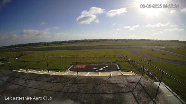 A view from a webcam at Leicestershire Aero Club shows a sunny day with a few scattered clouds in the sky. The scene includes a grassy area with a small helipad marked with a large white "H" on the ground. In the background, there is a paved runway and open green fields. The timestamp in the top right corner reads "02-06-2025 08:00:24." The bottom left corner has the text "Leicestershire Aero Club."