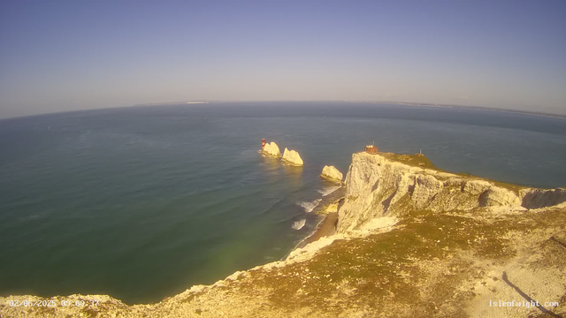 Aerial view of white chalk cliffs with patches of green grass, overlooking the ocean. There are striking white rocks, some with red and white lighthouses, emerging from the water. The sky is clear and blue, and the water appears calm with gentle waves. A small building is situated near the edge of the cliff. The timestamp in the bottom left corner shows "02/06/2025 09:00:37," and a website URL "isleofwight.com" is visible in the bottom right corner.