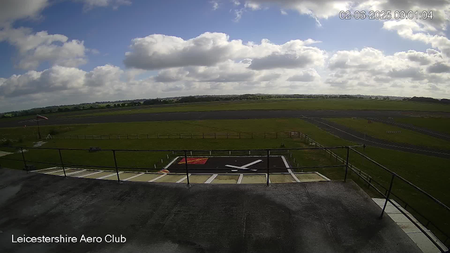 A view from a webcam at Leicestershire Aero Club showing a partly cloudy sky with a few large clouds and a bright blue sky. The aircraft landing area with a black and white markings is visible in the foreground. The surrounding green grass fields extend into the distance, with a runway and some small structures. A windsock is positioned on the left side of the image. The timestamp indicates the photo was taken on February 6, 2025, at 09:01:04.