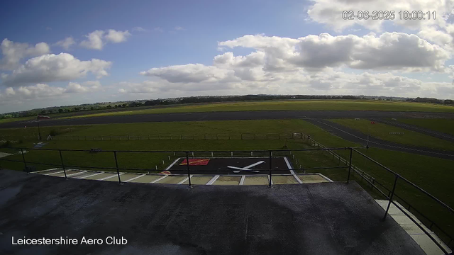 An outdoor scene at Leicesterhire Aero Club showing an airfield with a tarmac runway and a helicopter landing pad in the foreground. The sky is partly cloudy with white clouds and patches of blue. There are some hills and fields in the distance. The timestamp on the image is February 6, 2025 at 10:00:11.