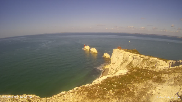 A view of a coastal landscape with white chalk cliffs and rock formations in the sea. There is a red and white lighthouse on one of the rocks, and a small building on top of the cliffs. The sea is calm and various sailboats are visible in the distance under a partly cloudy sky. The bottom left corner shows a timestamp, and the bottom right corner has a website URL.