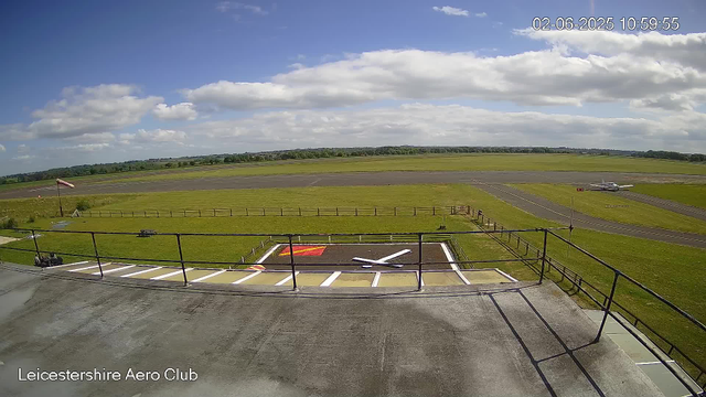 An outdoor view of an airfield on a bright day with partly cloudy skies. In the foreground, there is a rooftop helipad marked with a white "H" inside a square, and a small section with red and yellow markings. A black railing surrounds the helipad. To the left, a windsock is visible, indicating the wind direction. The runway extends across the middle of the image, with a small aircraft parked on the right side. The background consists of a vast grassy area and distant trees, with a few scattered clouds in the sky. The image includes a timestamp showing "02-06-2025 10:59:55" and a label "Leicestershire Aero Club" at the bottom left corner.