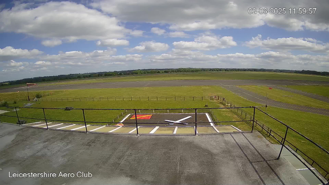 A view of the Leicestershire Aero Club airfield under a partly cloudy sky. The foreground shows a small platform with railings, marked with a red and yellow helicopter landing pad. Beyond the platform, there is a grassy area enclosed by a fence, and a windsock on a pole to the left. The runway stretches out in the distance, with a rural landscape and rolling hills in the background. The timestamp in the upper right corner indicates the date and time as February 6, 2025, at 11:59:57.