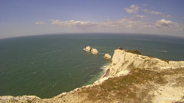 An aerial view of a coastal scene showing white chalk cliffs with a grassy top on the right side, extending into the sea. There are several large rocks jutting out of the water near the cliffs, with a small red and white lighthouse on one of the rocks. The sea is a muted greenish-blue, with boats creating white trails across the water. The sky is mostly clear with a few scattered clouds.