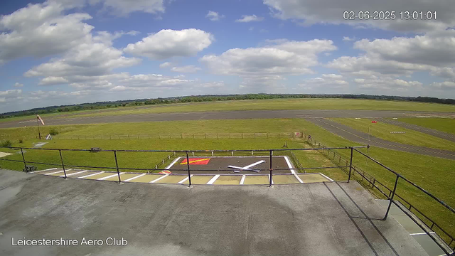 A view from a webcam at Leicestershire Aero Club showing an open grassy area with a small landing pad and a flagpole, overlooking a runway under a partly cloudy sky. The timestamp reads 02-06-2025 13:01:01.
