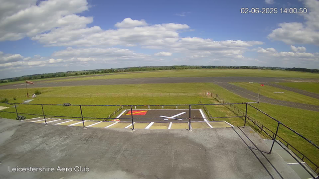 An outdoor view from a webcam at Leicestershire Aero Club showing a clear sky with scattered clouds. The image captures a grassy airfield with a runway extending into the distance. In the foreground, there is a helipad marked with a large white "H" and red and white painted markings. A small wind sock is visible near the helipad, and barriers surround the area. The landscape beyond the airfield is flat with green fields and distant trees on the horizon. The timestamp on the image indicates it was taken on February 6, 2025, at 14:00:50.