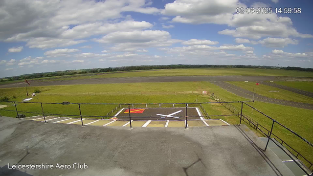 A view from a webcam at Leicestershire Aero Club shows a clear day with a partly cloudy sky. The foreground features a small helipad with white and red markings, surrounded by a black railing. Beyond the helipad, there are grassy areas and paved taxiways or runways stretching into the distance. In the top right corner, a timestamp reads "02-06-2025 14:59:58". The landscape is flat with some low hills or tree lines in the far background.