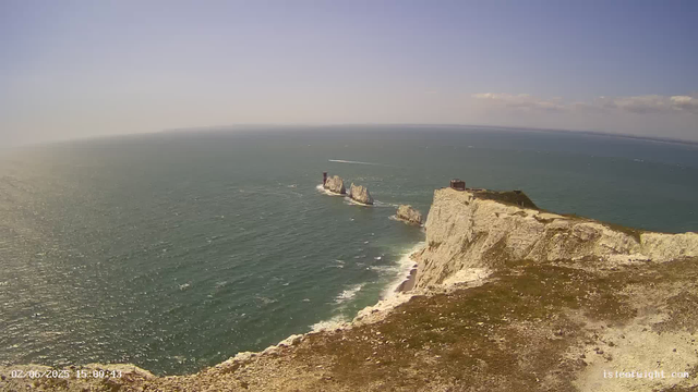 A view from a webcam showing white chalk cliffs overlooking the sea, with a small lighthouse and a building on top of the cliffs. The ocean below is calm with visible ripples, and several boats are moving in the distance. The sky is mostly clear with a few clouds near the horizon. The timestamp in the bottom left corner reads "02/06/2025 15:00:43".