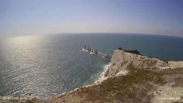 Aerial view of white chalk cliffs with greenery on top stretching along the coastline, with a small building near the edge. The sea below sparkles in the sunlight, with rocky stacks emerging from the water near the base of the cliffs. The sky is clear and blue.