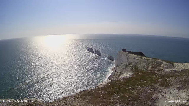 A view of tall white chalk cliffs meeting the ocean, with several large rocks or sea stacks near the shoreline. The sun is shining brightly, creating a sparkling reflection on the water. The sky is clear and blue, and a small building or structure is visible on top of the cliffs. The image has a timestamp of February 6, 2025, at 17:00:06.