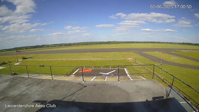 A view from a webcam showing a clear day at Leicestershire Aero Club. In the foreground, there is a small helipad with a white helicopter silhouette painted on it, surrounded by a safety railing. Beyond the helipad, there is a grassy area with a small flagpole flying a red flag. In the background, there are taxiways and runways extending into a flat landscape with green fields and a line of trees on the horizon. The sky is mostly blue with scattered white clouds. The date and time in the top right corner indicate it is June 2, 2025, at 5:00 PM.