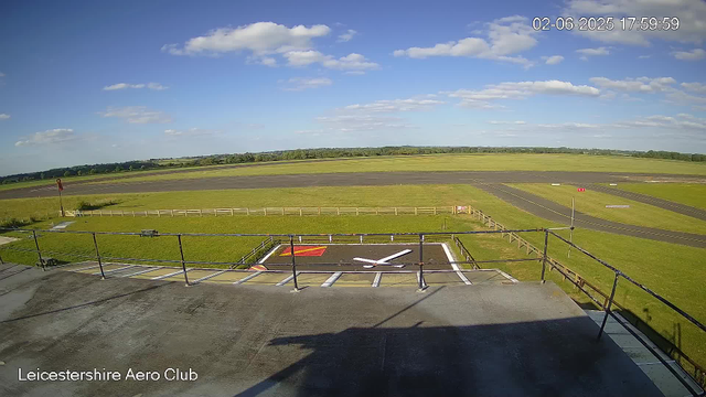 A view from a webcam at Leicestershire Aero Club shows a bright, sunny day with a blue sky and scattered white clouds. In the foreground, there is a metal balcony railing, and a small helipad with white markings and a red border is visible below. Beyond the helipad, a large grassy area is enclosed by a white fence, and an asphalt runway extends across the scene, with some markings and signs visible along its length. In the distance, there are open fields and a line of trees on the horizon. The timestamp indicates the date and time as June 2, 2025, at 17:59:59.
