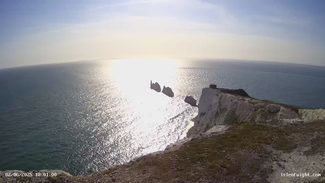 A view from a high cliff overlooking the ocean at sunset. The sky is partly cloudy with the sun low on the horizon, reflecting bright light on the water. In the distance, there are iconic white chalk stacks, including the famous Needle Stack and the Dlandogoras Rocks, rising from the sea. The foreground shows the edge of the cliff with rough, grassy, and rocky terrain. The scene is tranquil and scenic, capturing the natural beauty of the coastal landscape.