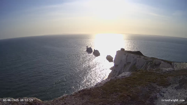 A scenic view of the ocean at sunset, with the sun casting a bright reflection on the water. White chalk cliffs extend into the sea, with a small building or lighthouse visible on one of the rocks in the distance. The foreground shows the edge of the chalky cliff with some patches of grass or moss. The timestamp on the lower left indicates the date and time as February 6, 2025, 6:59 PM, and the website is isleofwight.com.