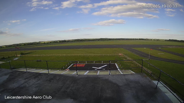 An empty runway at Leicester Aeroclub with a large helicopter landing pad in the foreground. There is a small red windsock on the left side, a wooden fence around the landing area, and a black tarmac runway extending into the distance under a partly cloudy sky. The image has a timestamp of June 2, 2025, at 19:00:33.