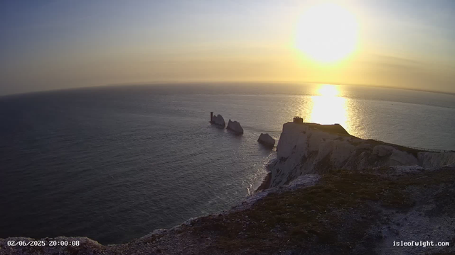 A scenic view of white chalk cliffs overlooking the ocean at sunset. The sun is low on the horizon, casting a warm glow and reflecting on the water. There are several large rock formations in the sea near the base of the cliffs. The image has a timestamp in the bottom left corner and a website address in the bottom right.